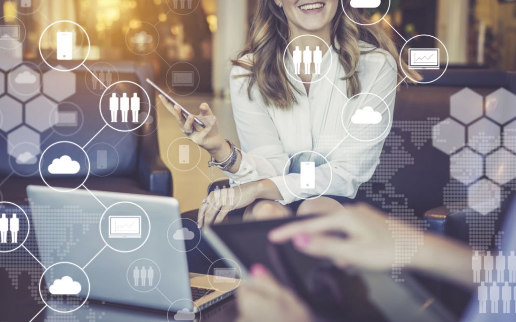 One-on-one meeting.Two businesswomen sitting at table and talking.On desk is laptop.Women have smartphone and digital tablet in their hands.In foreground are virtual icons with clouds,people,gadgets.